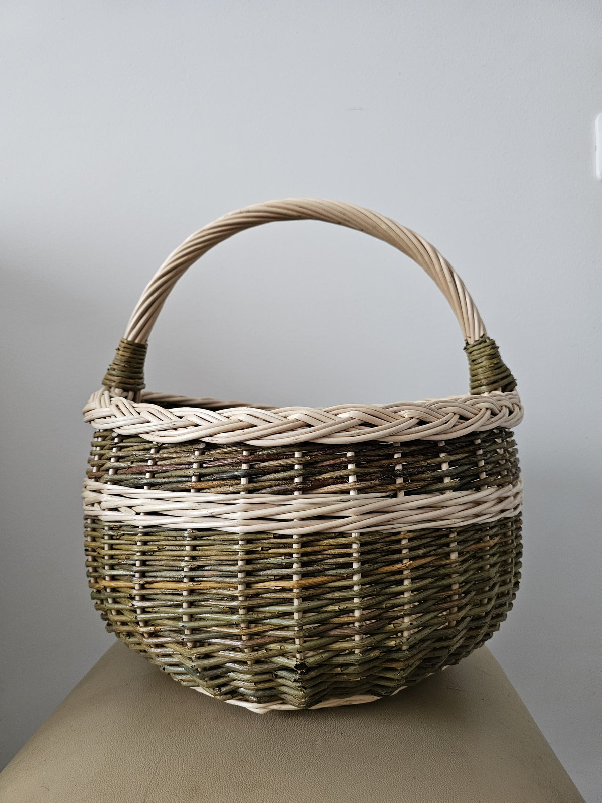 Round willow egg basket with a braided rim, arched handle and a woven band around the body, photographed against a plain grey wall
