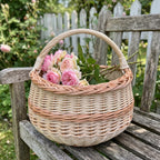 Round handwoven willow basket with handle holding a bunch of roses on a weathered wooden garden bench by a picket fence