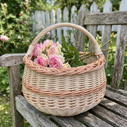Round handwoven willow basket with handle holding a bunch of roses on a weathered wooden garden bench by a picket fence