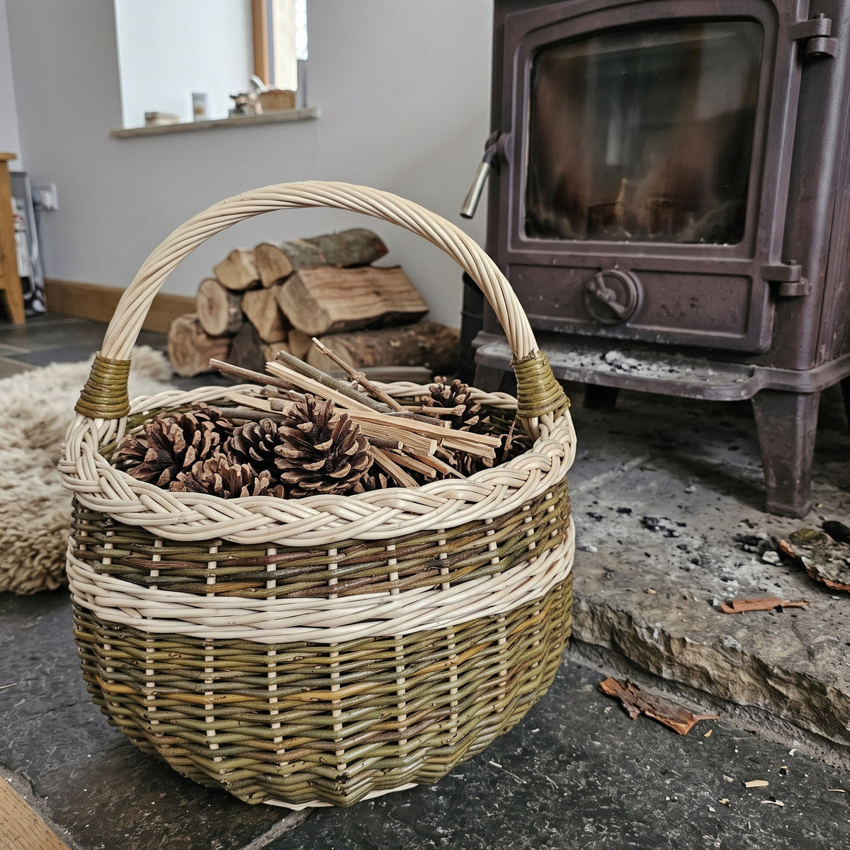 Round wicker basket with handle filled with pine cones and kindling beside a wood-burning stove and stacked firewood