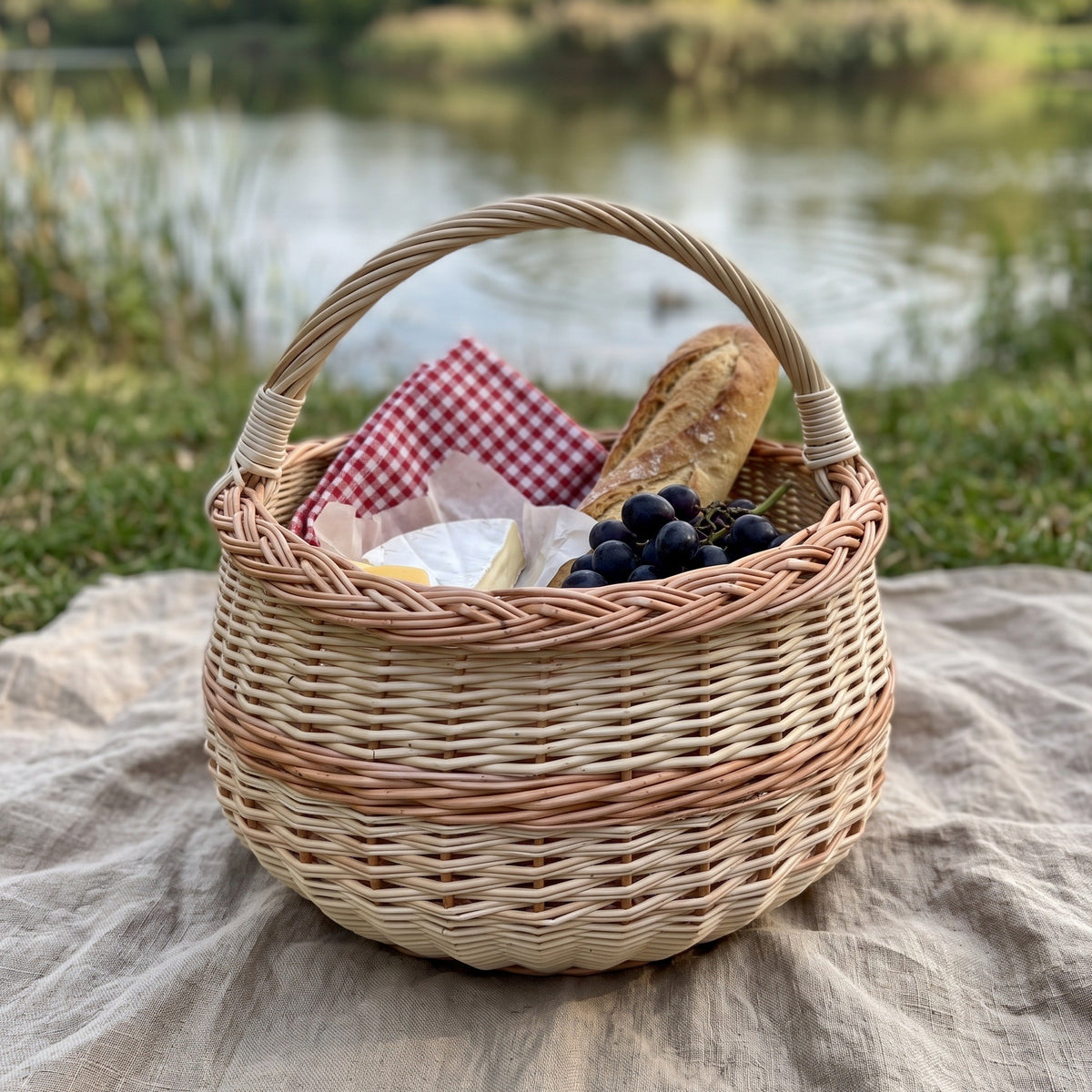 Round willow picnic basket with handle packed with a baguette, grapes and a checked napkin on a blanket by a lake