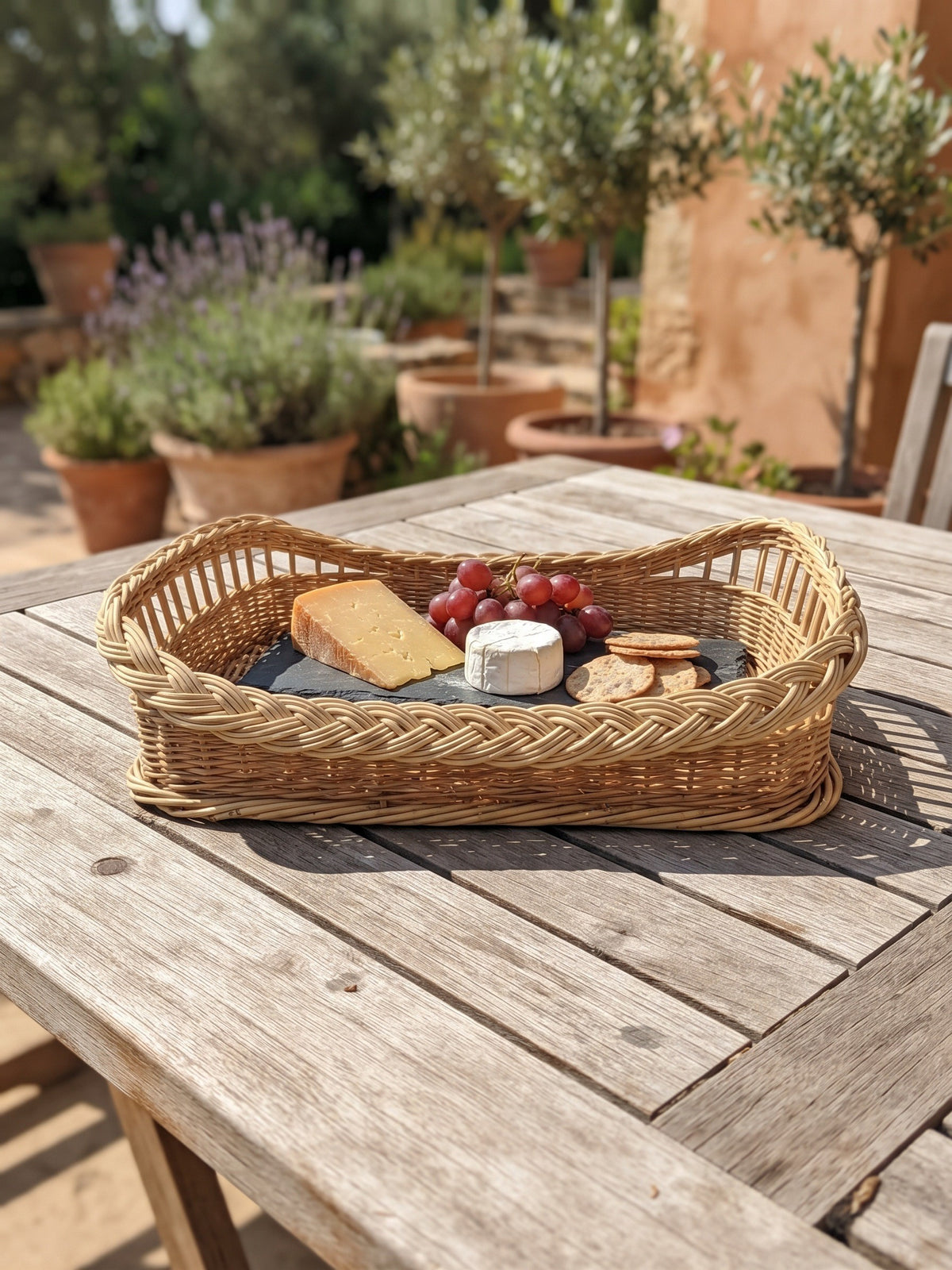 Rectangular wicker cheese tray with a wooden base holding cheese, crackers and grapes on a slate board on a sunny garden terrace table.