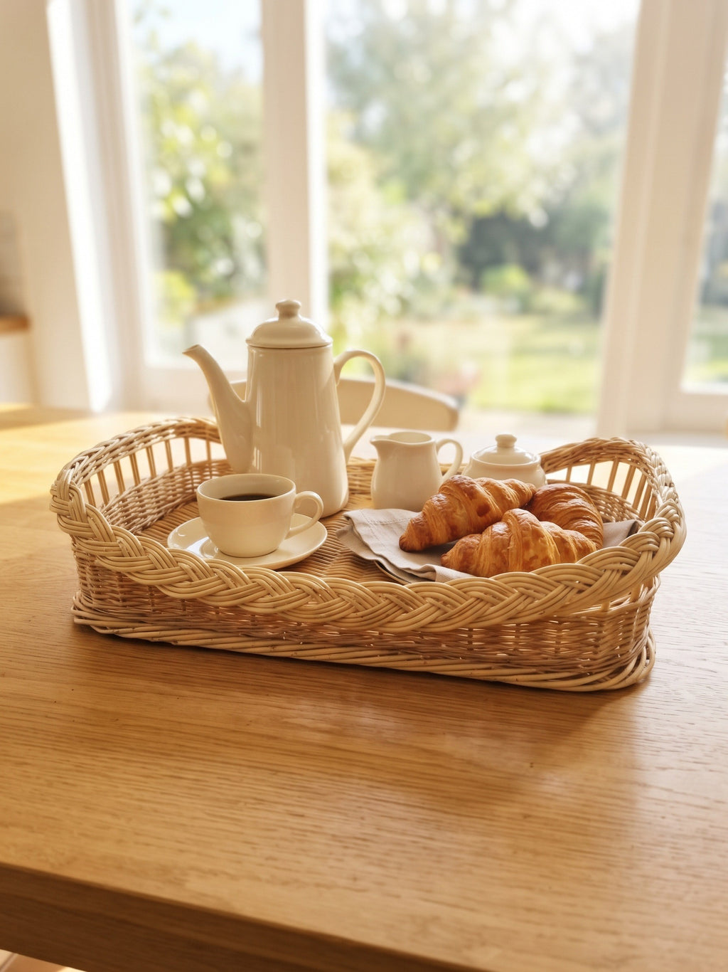 Willow tray with a braided rim holding a coffee pot, cups and croissants on a kitchen table in front of a bright garden window.