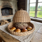Wicker bread cloche raised above its braided tray holding a rye loaf and bread rolls on a rustic table in front of a traditional brick bread oven.