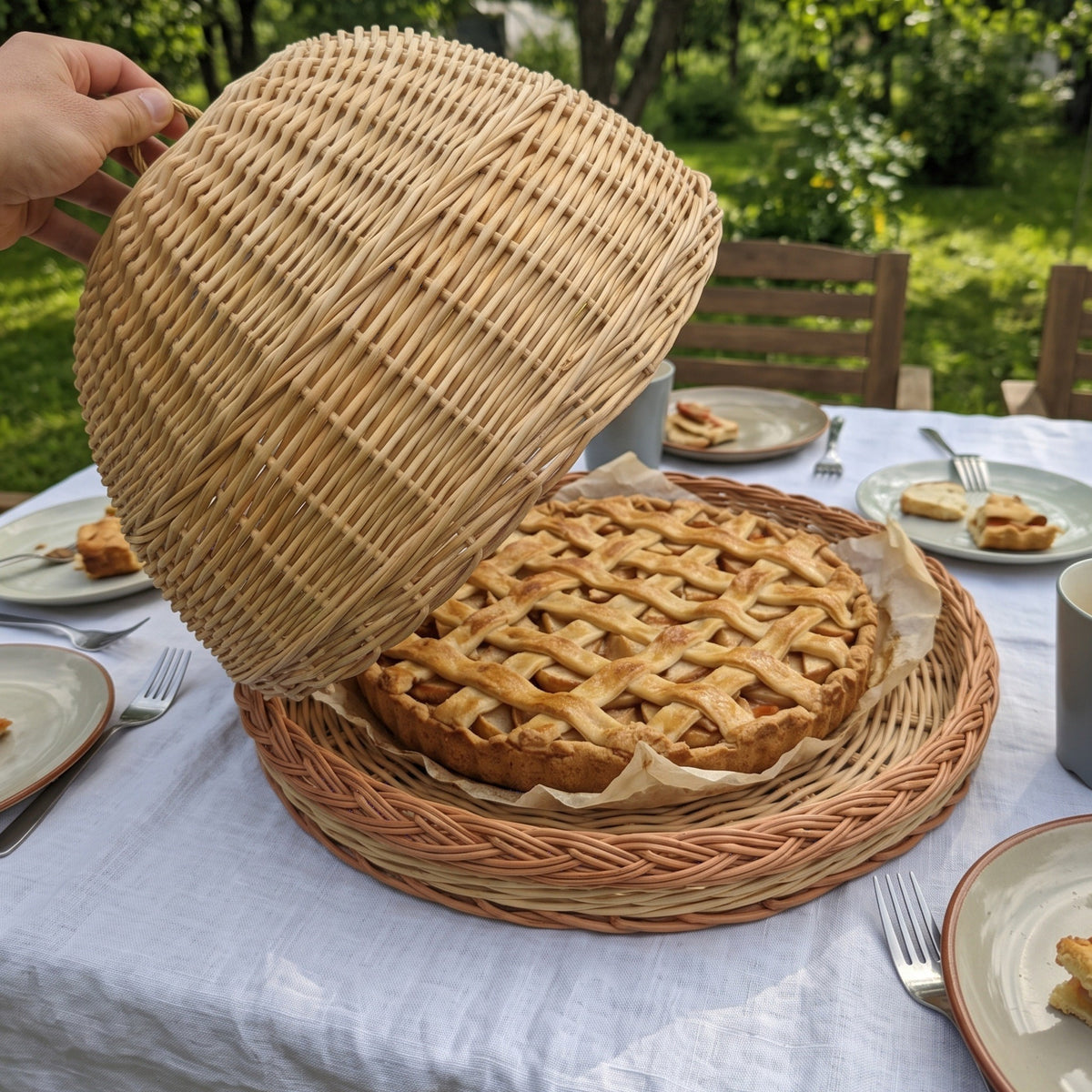 Wicker food cover lifted from its woven tray to reveal a lattice-top pie on a garden table set with plates and pastries for an outdoor lunch.