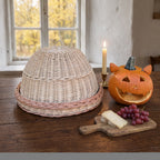 Handwoven wicker cloche with tray on a farmhouse table by a window, beside a carved pumpkin, a lit candle, cheese and grapes for an autumn evening.