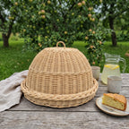 Willow food cover with tray on an outdoor wooden table in an apple orchard, next to a slice of sponge cake, a jug of lemonade and ceramic cups.