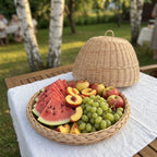 Wicker food dome placed behind its woven tray filled with watermelon slices, peaches, apples and grapes on a linen-covered garden table among birch trees.