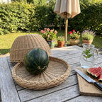 Willow cloche resting behind its round woven tray holding a whole watermelon on a patio table with potted flowers, a mint water jug and cut melon.