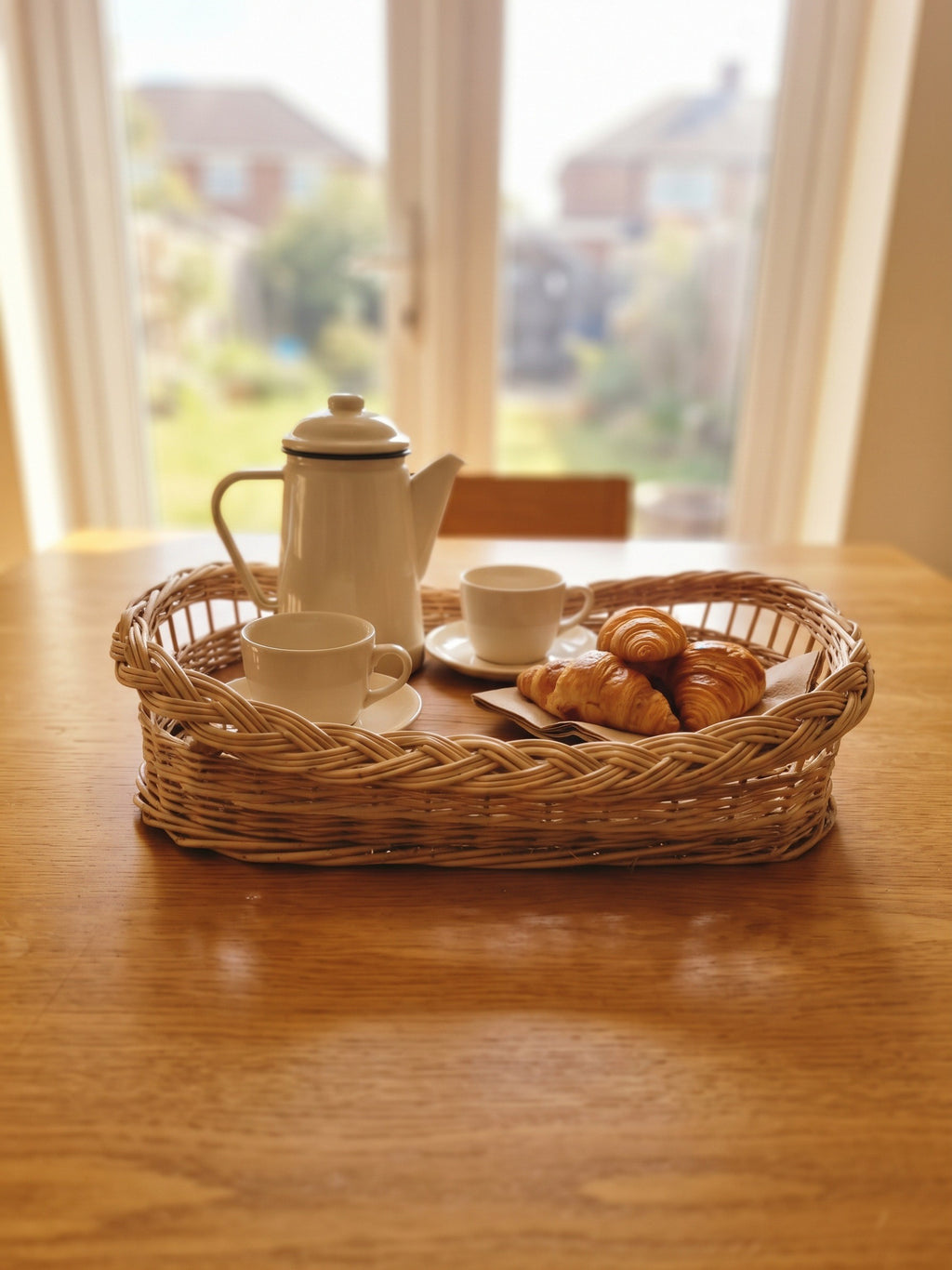Handwoven willow coffee tray with braided rim serving an enamel coffee pot, cups and croissants on a wooden dining table