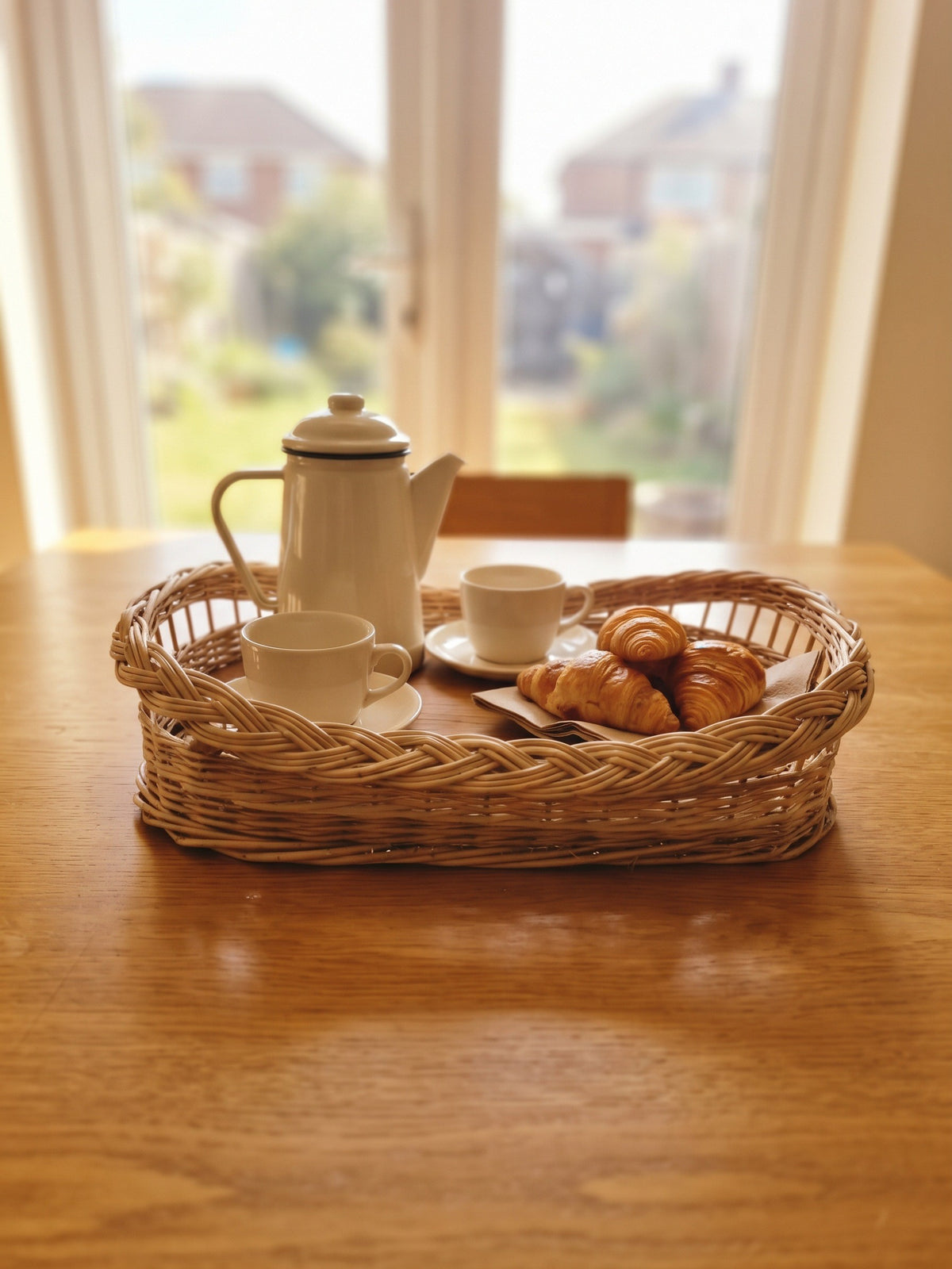 Handwoven willow coffee tray with braided rim serving an enamel coffee pot, cups and croissants on a wooden dining table