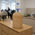 Wicker cooler sleeve hiding a 5 gallon water jug with a hand pump on an office kitchen counter, with paper cups and glasses nearby
