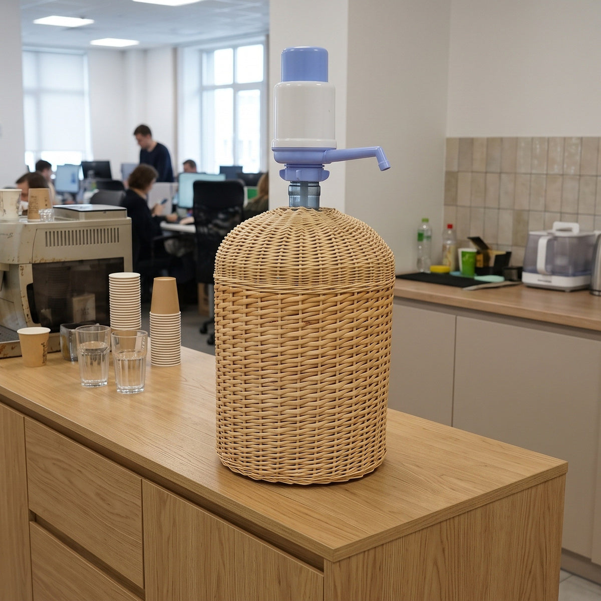 Wicker cooler sleeve hiding a 5 gallon water jug with a hand pump on an office kitchen counter, with paper cups and glasses nearby