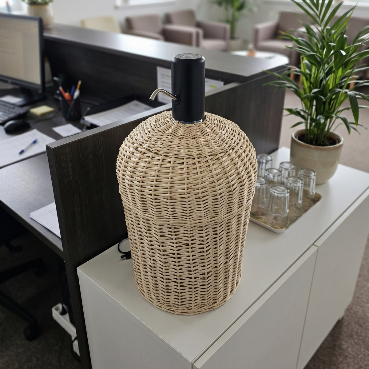 Wicker water cooler cover on a 5 gallon bottle topped with an electric pump, set on a white cabinet in a modern open-plan office