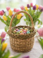 Small handwoven Easter basket with handle filled with pastel dyed eggs on moss, surrounded by vases of spring tulips on a table.