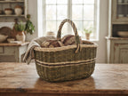 Handwoven oval willow market basket with a twisted handle on a rustic kitchen table, holding fresh loaves of bread under a linen cloth