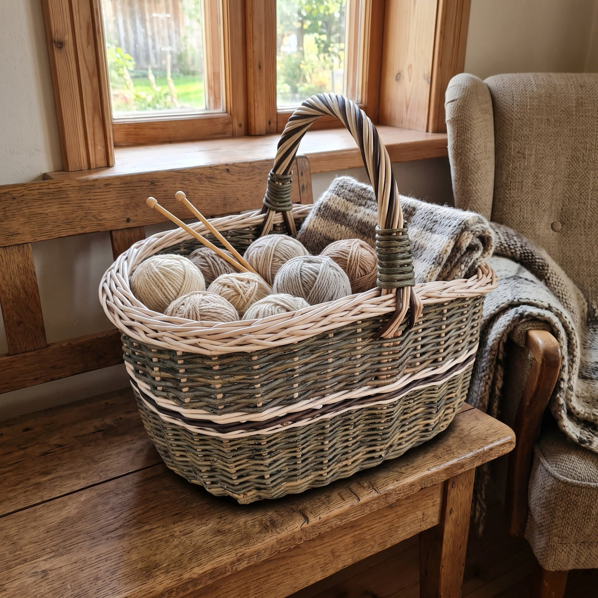 Oval wicker knitting basket holding balls of yarn and knitting needles on a wooden bench by a window, with a wool blanket draped beside it