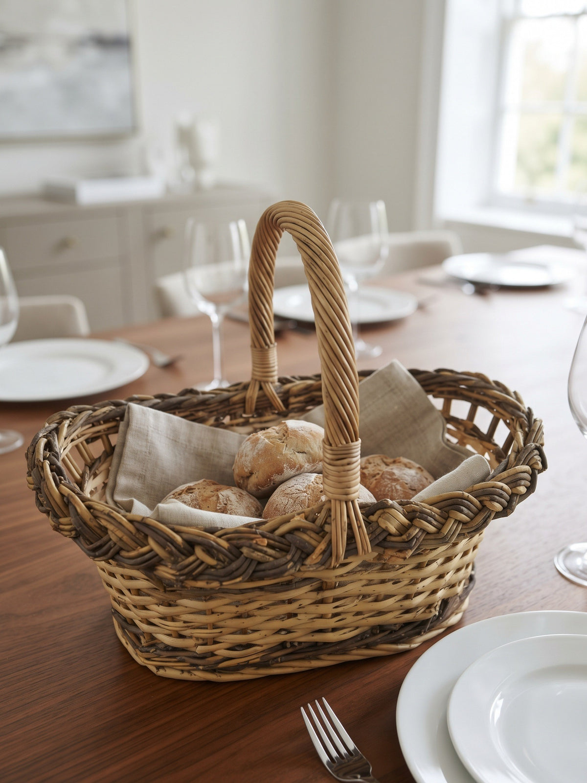 Handwoven willow bread basket with a braided rim holding bread rolls on a linen napkin on a dining table set with plates and glasses.