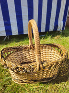Oval wicker bread basket with a tall arched handle and braided rim standing on the grass in front of a striped market tent.