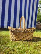 Side view of a small handwoven willow bread basket with a braided rim and curved handle on sunny grass by a striped tent.