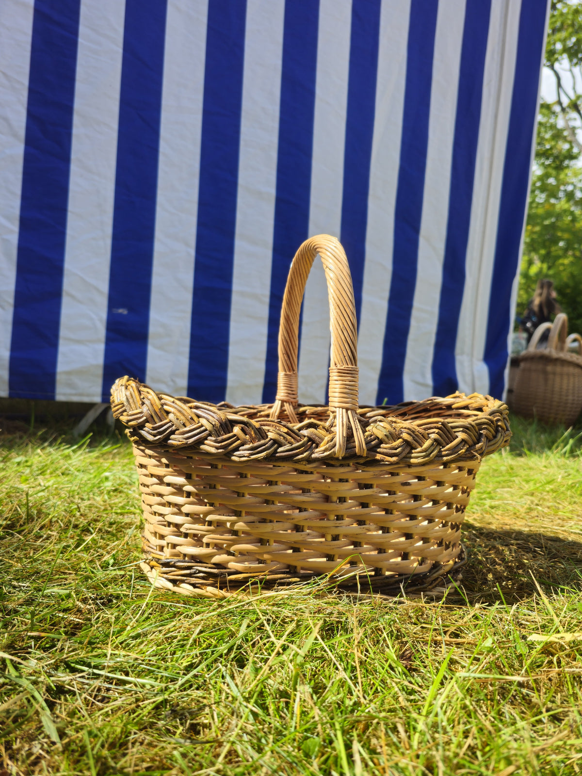 Side view of a small handwoven willow bread basket with a braided rim and curved handle on sunny grass by a striped tent.