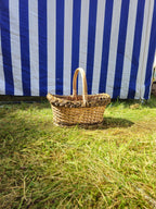 Small wicker bread basket with handle and decorative braided border standing on the lawn in front of a striped market tent.