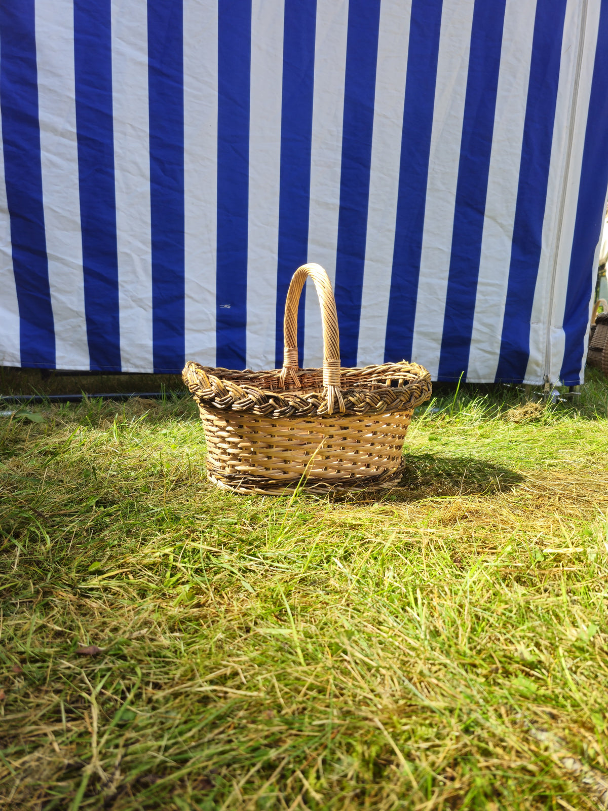 Small wicker bread basket with handle and decorative braided border standing on the lawn in front of a striped market tent.
