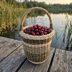 Tall willow berry picking basket full of ripe cherries, with a braided rim and arched handle, resting on a wooden jetty by a calm lake.