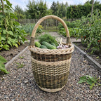 Handwoven tall wicker basket with handle carrying cucumbers, fresh dill and garlic along a gravel path in a vegetable garden.