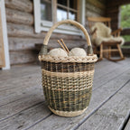 Tall wicker knitting basket with handle holding balls of yarn and wooden knitting needles on the porch of a log cottage.