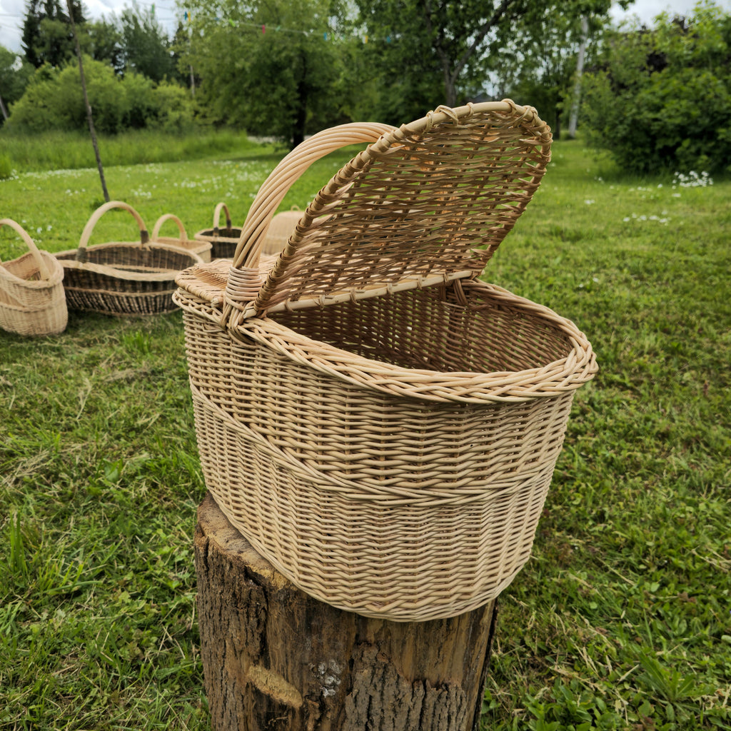 Wicker picnic hamper with a lid raised on a tree stump in a garden, with other handwoven willow baskets standing on the grass behind it
