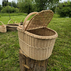 Wicker picnic hamper with a lid raised on a tree stump in a garden, with other handwoven willow baskets standing on the grass behind it