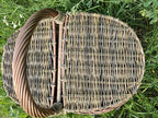 Top view of the oval wicker hamper on the grass, its two hinged lids meeting neatly under the sturdy twisted central handle.