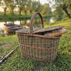 Wicker hamper with a double lid and a loaf of bread inside, standing on a riverbank at sunset beside a fishing rod and a boat.
