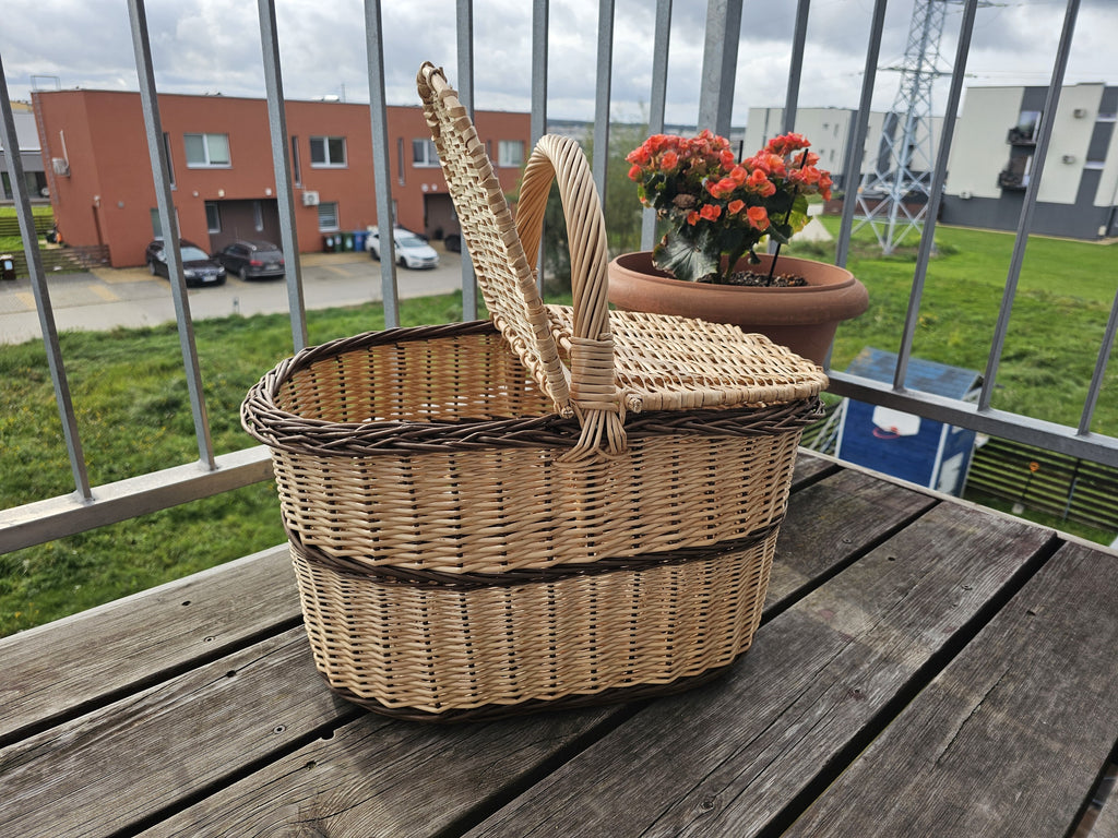 Oval wicker picnic hamper with one lid lifted open, showing the deep interior, on a wooden balcony table.