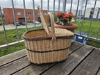 Oval wicker picnic hamper with one lid lifted open, showing the deep interior, on a wooden balcony table.