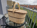 Handwoven willow picnic hamper with two lids and a centre handle on a round glass table on a balcony.