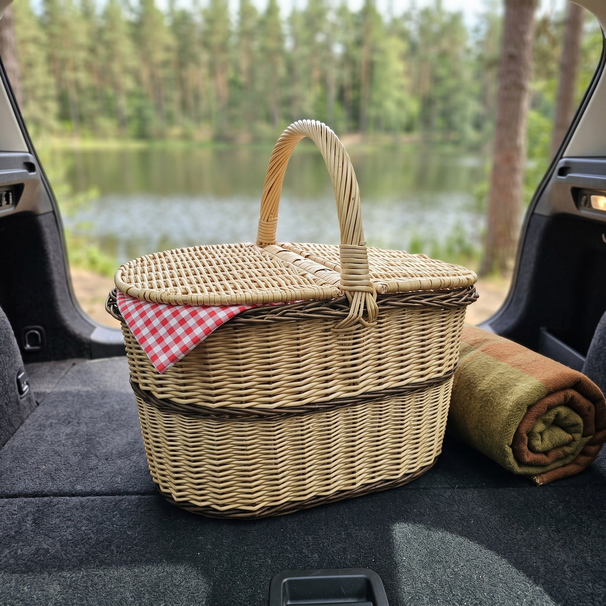 Oval wicker picnic hamper with lid in the open boot of a car by a lake with a checked napkin and a rolled blanket.