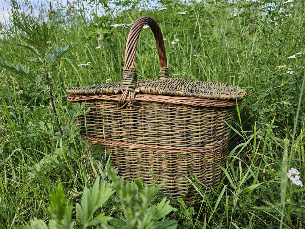 Willow picnic basket with hinged lids and an arched handle standing in a meadow, showing the interlaced weave over a sturdy base.