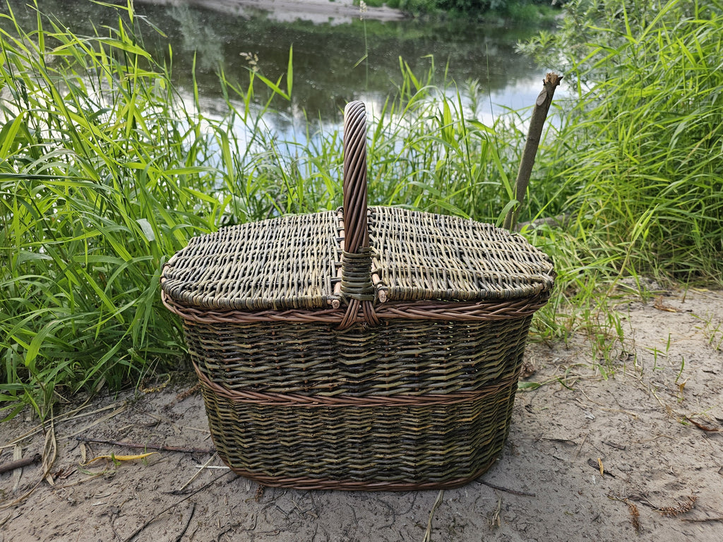 Oval willow picnic hamper with lid and centre handle standing on sand by a lake among tall green grass.