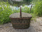 Oval willow picnic hamper with lid and centre handle standing on sand by a lake among tall green grass.