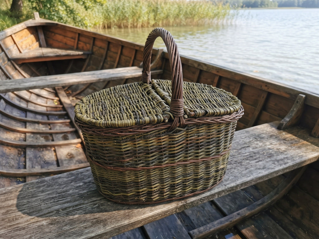 Large picnic basket with a double lid, handwoven from unpeeled willow, resting on the bench of a wooden rowing boat moored on a lake.