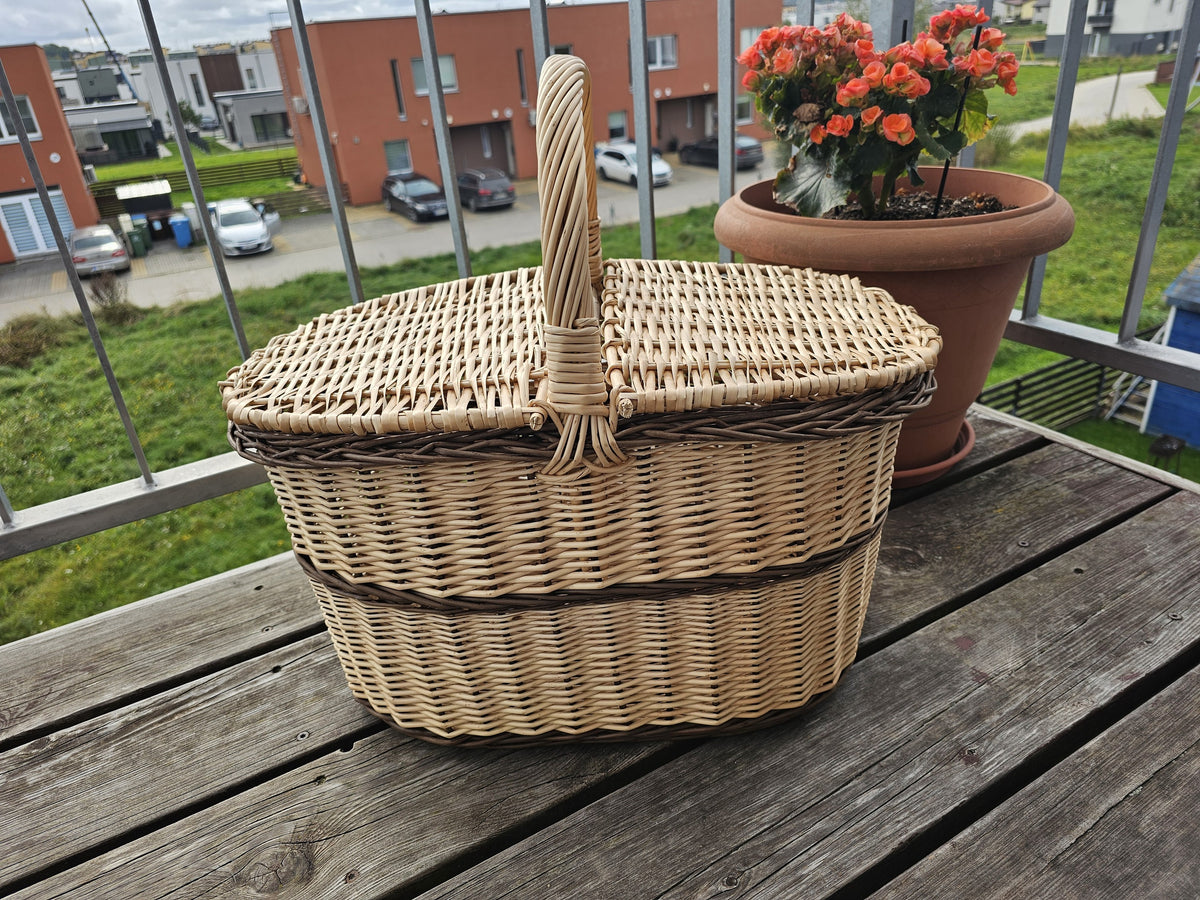Oval wicker picnic hamper with two hinged lids and a centre handle on a wooden balcony table beside a pot of flowers.