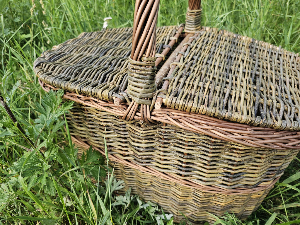 Close-up of the handwoven willow picnic basket with a strong arched handle and twin lids that open from the centre, set on the grass.