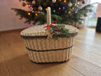 Oval wicker hamper with lid decorated with a red bow and fir sprigs as a Christmas gift basket in front of a decorated tree.