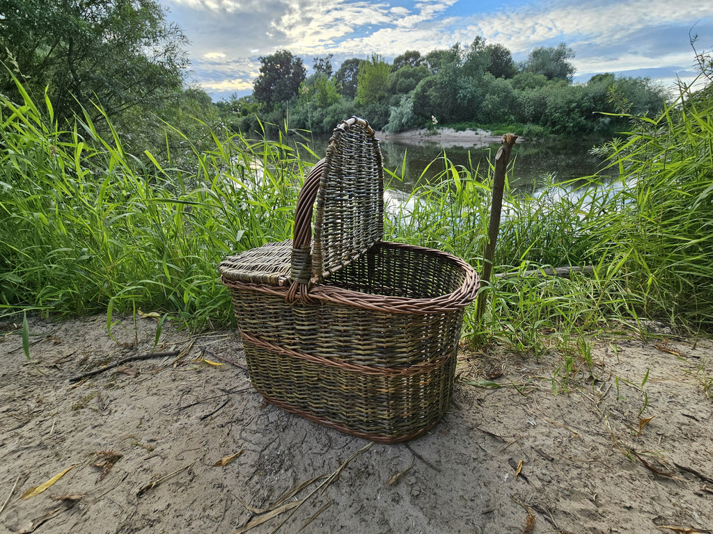 Large willow picnic hamper with an arched handle and one lid propped open on a sandy riverbank, showing the hollow interior and textured weave.