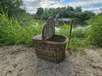 Large willow picnic hamper with an arched handle and one lid propped open on a sandy riverbank, showing the hollow interior and textured weave.