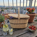 Handwoven willow picnic hamper with lid holding a baguette and lavender, set with lemonade and strawberries on a balcony table.
