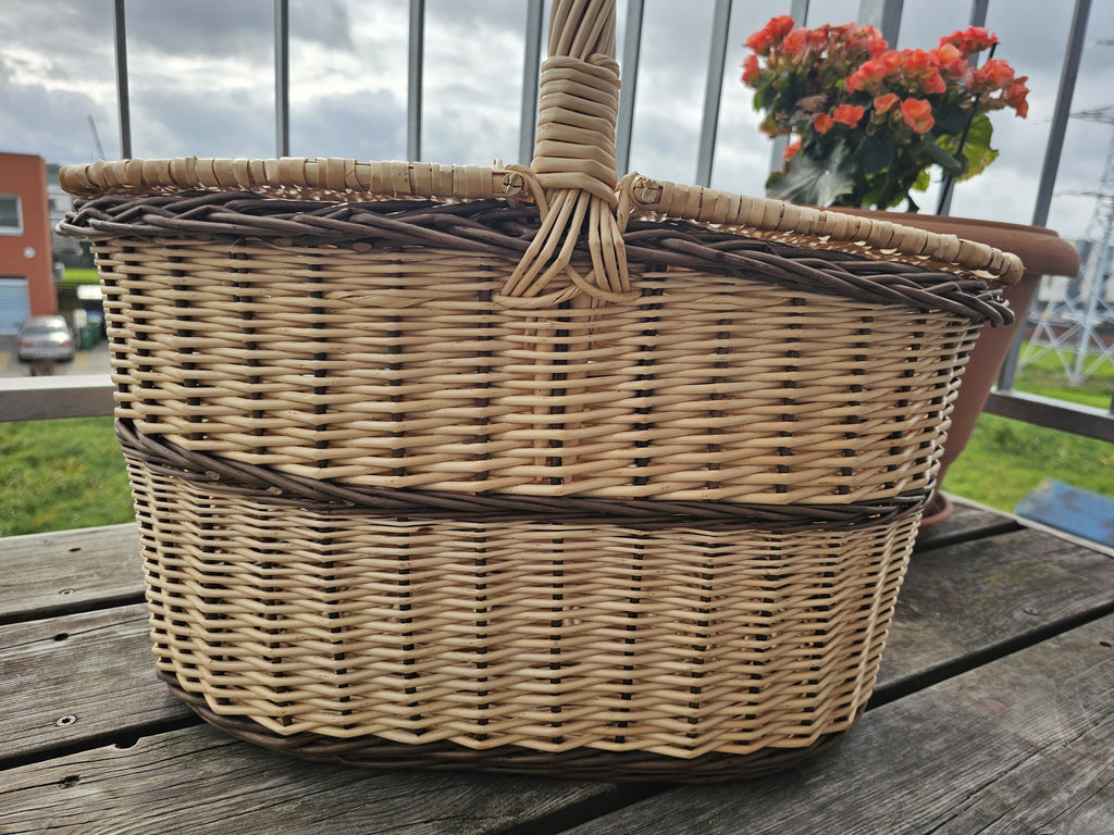 Low side view of the oval handwoven wicker picnic hamper with lid, showing the woven bands, rim and centre handle on a balcony.