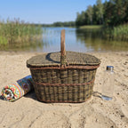 Large willow picnic hamper with a central handle on a sandy lake beach beside a rolled blanket and a glass water bottle.